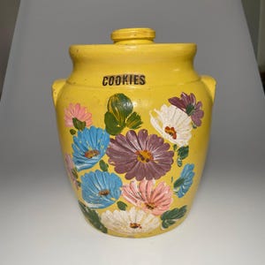 May include: A yellow ceramic cookie jar with a matching lid and two side handles. The front features hand-painted floral designs in blue, purple, pink, and white with green leaves. The word COOKIES is printed in black letters near the top rim.