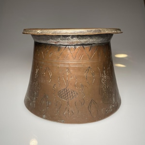 May include: A copper-colored metal container with a wide base and a narrower top. The container features etched geometric and floral designs. The rim has a silver-colored band. The container is likely an antique or vintage item.