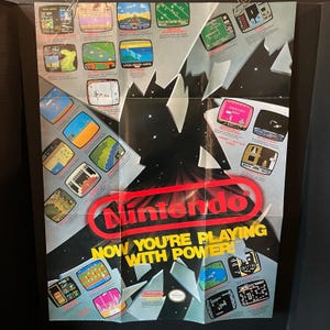 May include: A vintage Nintendo Entertainment System promotional poster featuring a central red Nintendo logo and the text "NOW YOU'RE PLAYING WITH POWER!" surrounded by numerous small screenshots of various classic video games on a dark background.