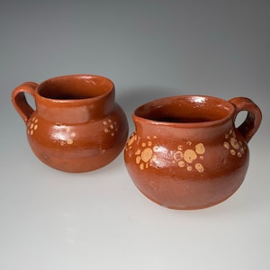 May include: Two rustic, brown ceramic mugs with handles. Each mug has a rounded body and is decorated with small, light-colored polka dots. The mugs are likely handmade and suitable for serving beverages.