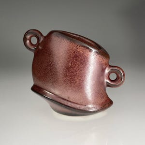 May include: A handmade ceramic mug with a unique, asymmetrical design. The mug is a deep reddish-brown color with a speckled texture and two loop handles. The rim is slightly angled, adding to its artistic appeal.