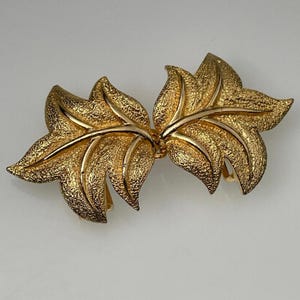May include: A gold-toned leaf-shaped brooch. The brooch features two symmetrical leaf clusters, each with textured detailing and a central vein. The leaves are connected by a gold bar. The brooch is likely vintage.