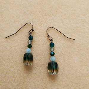 Puede incluir: Un par de pendientes colgantes con un esquema de color verde y azul. Los pendientes presentan un gran cristal facetado en forma de lágrima con cristales más pequeños y cuentas por encima.