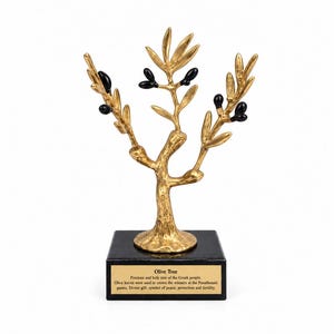 May include: A gold-colored metal olive tree sculpture with black olives attached to the branches. The tree stands on a square black base featuring a gold plaque with the text: Olive Tree, Precious and holy tree of the Greek people.