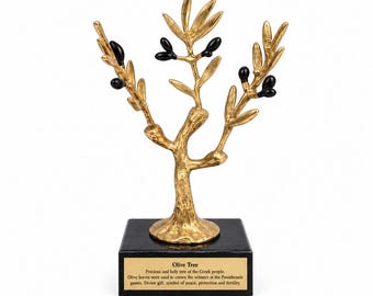 May include: A gold-colored metal olive tree sculpture with black olives attached to the branches. The tree stands on a square black base featuring a gold plaque with the text: Olive Tree, Precious and holy tree of the Greek people.