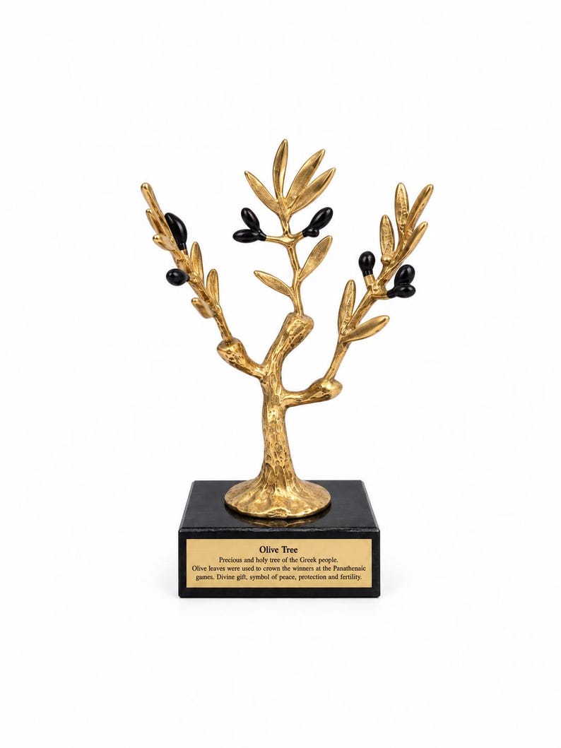 May include: A gold-colored metal olive tree sculpture with black olives attached to the branches. The tree stands on a square black base featuring a gold plaque with the text: Olive Tree, Precious and holy tree of the Greek people.
