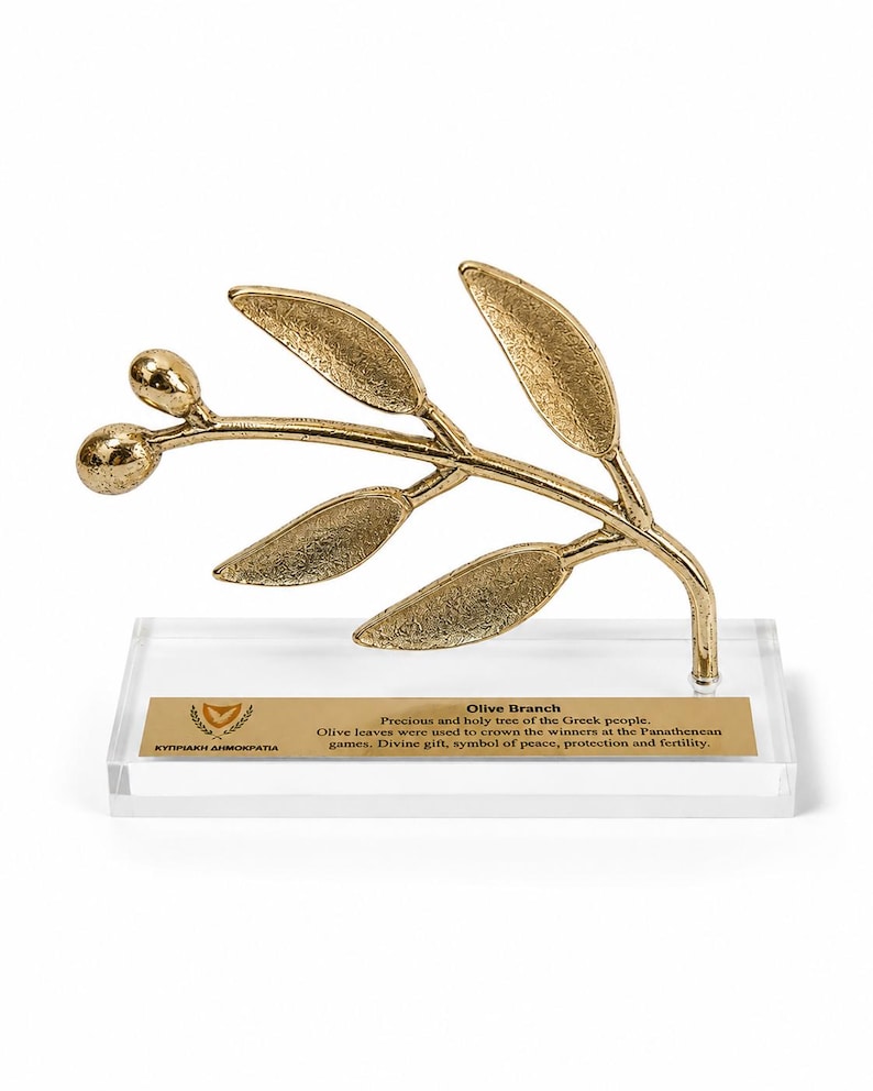 May include: A gold-colored metal olive branch sculpture with two olives and five leaves, mounted on a clear rectangular acrylic base. A gold plaque on the base features the text: Olive Branch, Precious and holy tree of the Greek people.