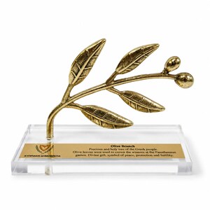 May include: A gold-toned metal olive branch sculpture with five leaves and two olives, mounted on a clear rectangular acrylic base. A gold plaque on the base features the text: Olive Branch, Precious and holy tree of the Greek people.