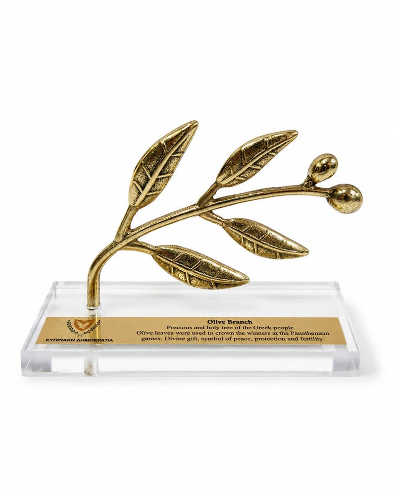May include: A gold-toned metal olive branch sculpture with five leaves and two olives, mounted on a clear rectangular acrylic base. A gold plaque on the base features the text: Olive Branch, Precious and holy tree of the Greek people.