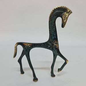 May include: A stylised metal horse figurine with a dark, oxidised finish and polished gold-toned highlights on the mane, face, and hooves. The body features engraved floral patterns and geometric motifs along the neck and back.