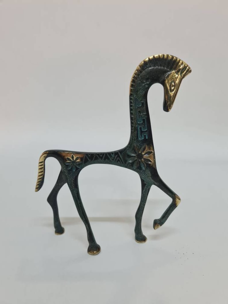 May include: A stylised metal horse figurine with a dark, oxidised finish and polished gold-toned highlights on the mane, face, and hooves. The body features engraved floral patterns and geometric motifs along the neck and back.