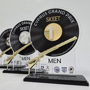 Puede incluir: Tres trofeos para la competición de Skeet del Gran Premio de Chipre. Cada trofeo presenta un disco negro con detalles dorados, un rifle dorado y una base negra. El trofeo del primer puesto tiene un centro dorado.