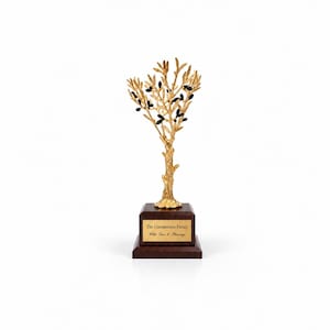 May include: A gold-colored metal olive tree sculpture with black oval-shaped olives attached to the branches. The tree is mounted on a square, dark brown wooden base featuring a gold-colored plaque with the text The Constantinou Family With Love & Blessings.