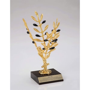 May include: A gold olive tree sculpture with black olives on a wooden base. The base has a plaque with the text "Olive Tree:  Symbol of peace and fertility, the olive tree is one of the oldest cultivated trees in the world. Olive branches were used as a symbol of peace, prosperity and life in ancient Greece.  The olive tree is a symbol of peace, prosperity and life."