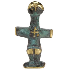 Może przedstawiać: Mała, zielona rzeźba z brązu przedstawiająca stylizowaną postać ludzką z krzyżem na piersi. Postać ma okrągłą głowę i wyciągnięte ramiona i stoi na małej podstawie.