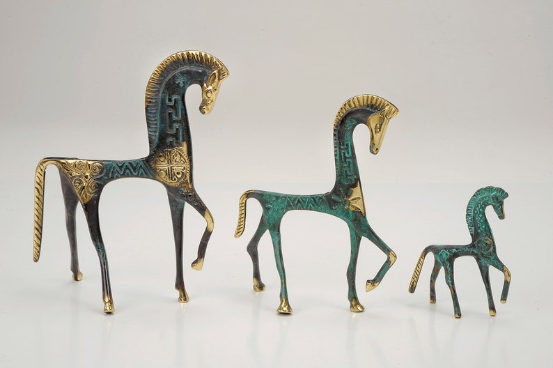 May include: Three horse figurines of varying sizes crafted from metal with a dark green patina and polished gold-tone accents. Each horse features a stylised mane, a slender body, and intricate geometric patterns engraved along the neck and torso.