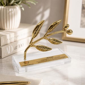 May include: A gold-colored metal olive branch sculpture featuring detailed leaves and two small olives, mounted on a clear rectangular acrylic base. A gold plaque on the base displays the text Olive Branch and Kypriaki Dimokratia with a small emblem.