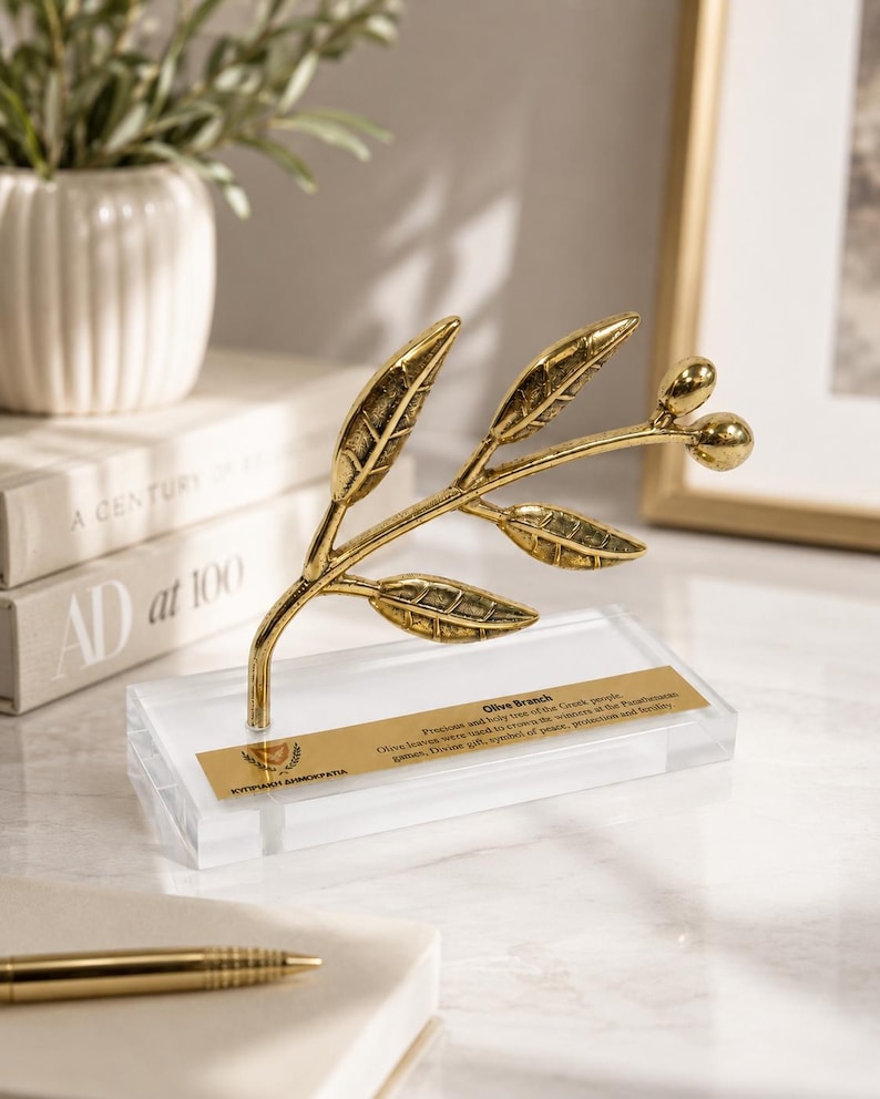 May include: A gold-colored metal olive branch sculpture featuring detailed leaves and two small olives, mounted on a clear rectangular acrylic base. A gold plaque on the base displays the text Olive Branch and Kypriaki Dimokratia with a small emblem.