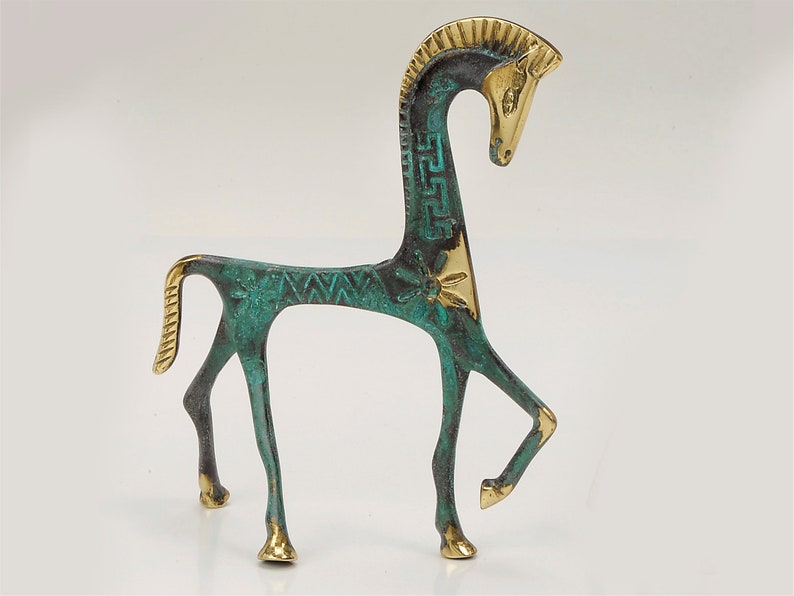 May include: A stylised horse figurine crafted from metal, featuring a combination of polished gold-tone accents and a textured, oxidised green patina. The design includes geometric Greek key patterns and floral motifs along the neck and body.
