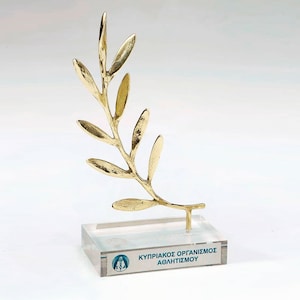 May include: A gold-colored metal olive branch sculpture mounted on a clear rectangular acrylic base. The base features a blue circular logo and the text "ΚΥΠΡΙΑΚΟΣ ΟΡΓΑΝΙΣΜΟΣ ΑΘΛΗΤΙΣΜΟΥ" printed in blue on the front face of the transparent material.