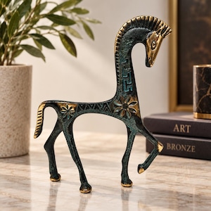 May include: A stylised horse figurine crafted from metal with a dark, oxidised green patina and polished gold-toned accents. The sculpture features intricate geometric patterns, floral motifs, and a Greek key design along its elongated neck.