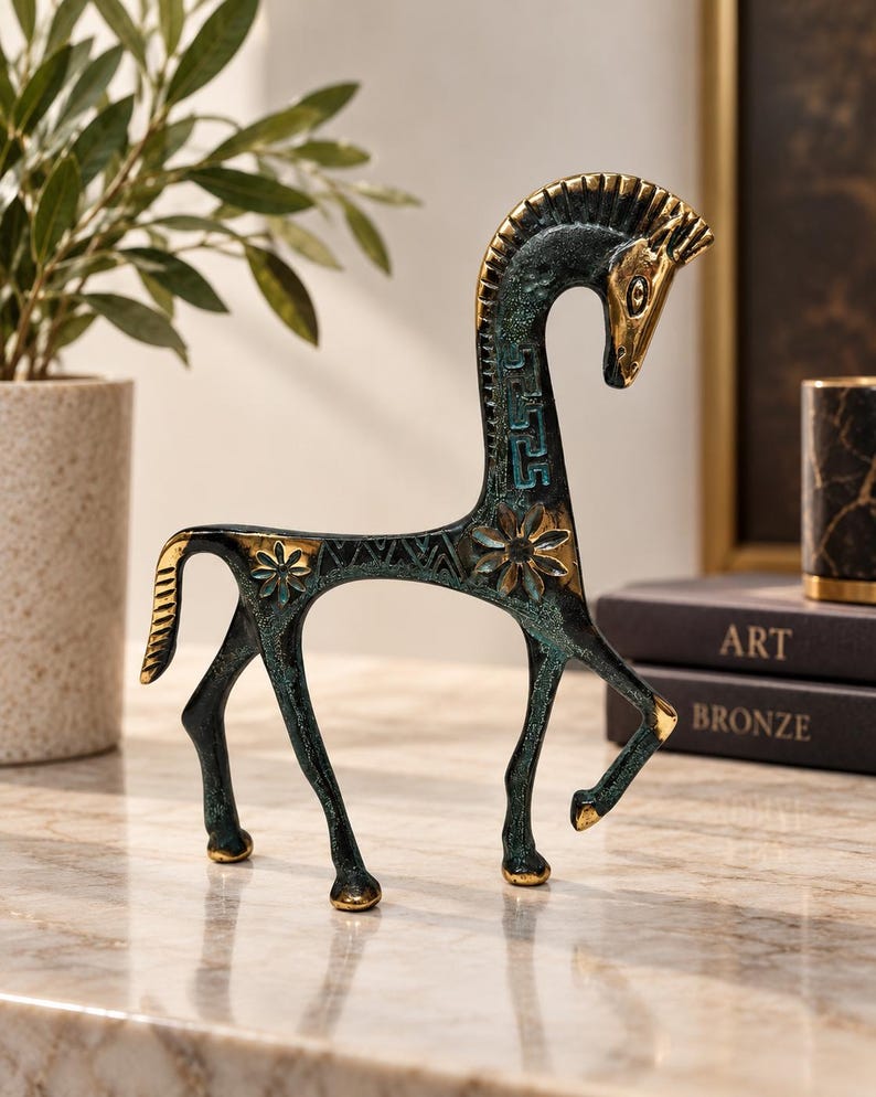 May include: A stylised horse figurine crafted from metal with a dark, oxidised green patina and polished gold-toned accents. The sculpture features intricate geometric patterns, floral motifs, and a Greek key design along its elongated neck.