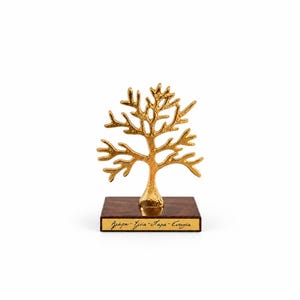 May include: A gold-colored metal tree sculpture with a textured finish, mounted on a rectangular dark brown wooden base. A small gold plaque on the front of the base features the Greek text: Agapi - Ygeia - Chara - Eftychia in black script.