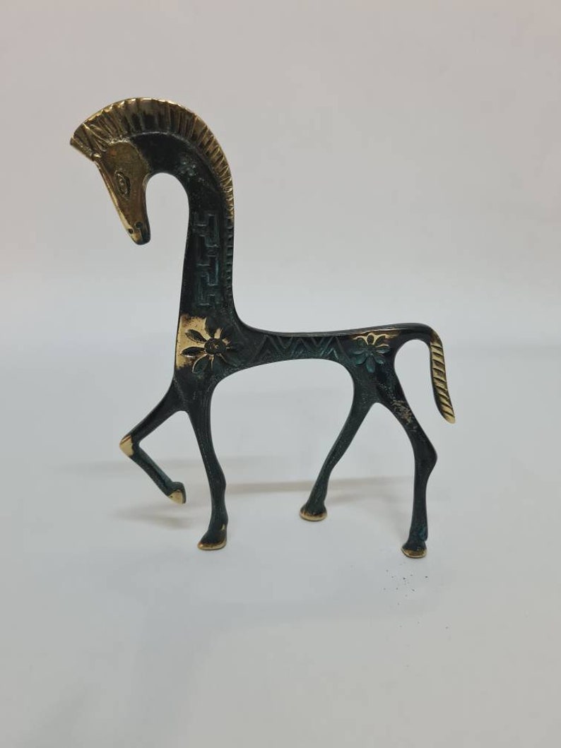 May include: A stylised metal horse figurine featuring a dark, oxidised finish with polished gold-tone accents on the mane, face, and tail. The body is decorated with engraved floral patterns and geometric lines, standing on four slender legs.