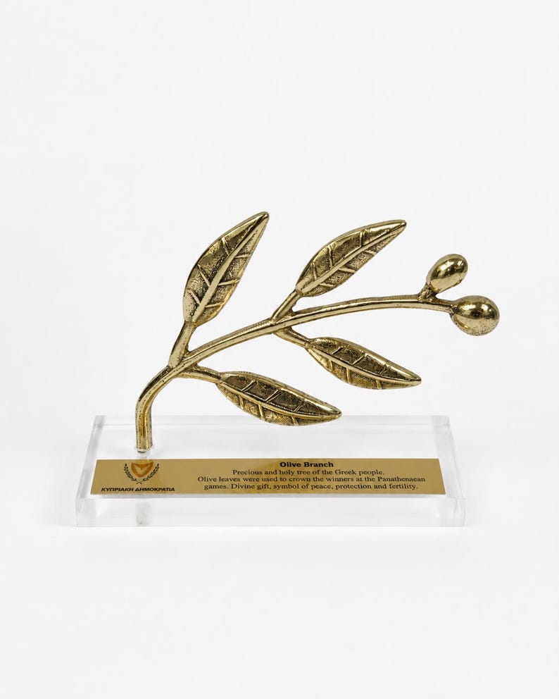 May include: A gold-toned metal olive branch sculpture with textured leaves and two small olives, mounted on a clear rectangular acrylic base. A gold plaque on the base features the text Olive Branch, the Cyprus emblem, and descriptive text.