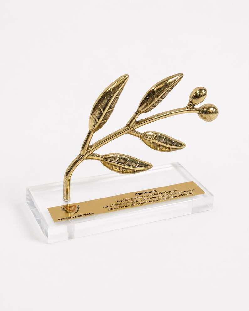 May include: A gold-toned metal olive branch sculpture featuring detailed leaves and two olives, mounted on a clear rectangular acrylic base. A gold plaque on the base includes the text Olive Branch, a description of its history, and the text KY&Pi;PIAKH &Delta;HMOKPATIA.