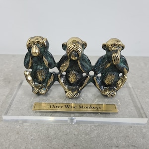 May include: Three gold-painted monkeys with green accents, each covering a different part of their face. The monkeys are on a clear acrylic base with a gold plaque that reads "Three Wise Monkeys".