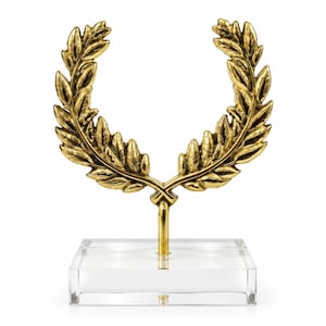 May include: A gold-colored metal laurel wreath sculpture mounted on a clear, rectangular acrylic base. The wreath features detailed, textured leaves arranged in a symmetrical, open-top circular shape, supported by a central vertical metal rod.