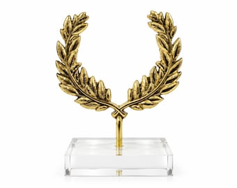May include: A gold-colored metal laurel wreath sculpture mounted on a clear, rectangular acrylic base. The wreath features detailed, textured leaves arranged in a symmetrical, open-top circular shape, supported by a central vertical metal rod.