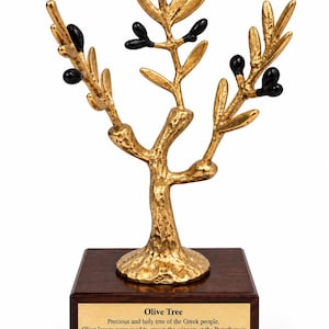 May include: A gold-colored metal olive tree sculpture with black olives and textured branches, mounted on a square dark brown wooden base. A gold plaque on the front reads: Olive Tree, Precious and holy tree of the Greek people. Olive leaves were used to crown the winners at the Panathenaic games. Divine gift, symbol of peace, protection and fertility.