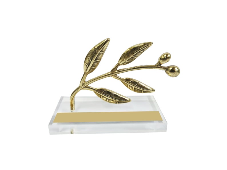 May include: A gold-colored metal olive branch sculpture featuring detailed leaves and two small olives, mounted on a clear rectangular acrylic base that includes a blank, rectangular gold-colored plaque on the front surface.