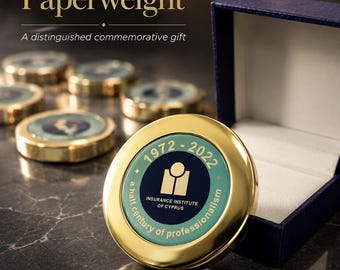 May include: A round gold-toned metal paperweight featuring a teal and dark blue center inlay. The text reads 1972-2022, Insurance Institute of Cyprus, and a half century of professionalism. It is presented in a dark blue open gift box.