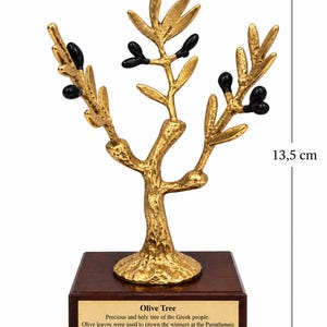 May include: A gold-colored olive tree sculpture with black olives, mounted on a square wooden base. A plaque on the base reads: Olive Tree, Precious and holy tree of the Greek people. Olive leaves were used to crown the winners at the Panathenaic games, Divine gift, symbol of peace, protection and fertility. Height is 13.5 cm.