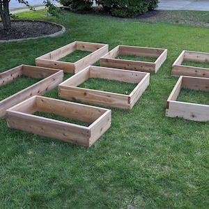 May include: Eight unfinished wooden raised garden beds of varying sizes are arranged on a green lawn. The rectangular beds are constructed from light-colored wood and are open at the top and bottom, ready for planting.