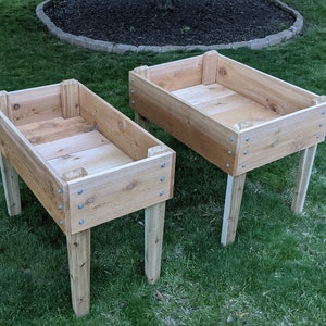 May include: Two rectangular wooden raised garden beds with four legs each. The beds are made of light-colored wood and are assembled with screws. They are placed on a green grassy lawn.