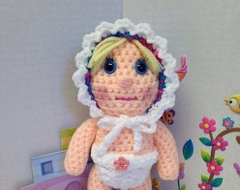 May include: A crocheted doll with blonde hair, blue eyes, and a pink and white dress. The doll is wearing blue and pink shoes and has a white crocheted bonnet.