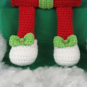 Puede incluir: Una mu&ntilde;eca de elfo de crochet con ropa roja y verde y zapatos blancos. El elfo est&aacute; sentado en una superficie verde con relleno blanco alrededor.