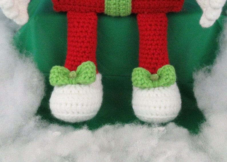 Puede incluir: Una mu&ntilde;eca de elfo de crochet con ropa roja y verde y zapatos blancos. El elfo est&aacute; sentado en una superficie verde con relleno blanco alrededor.
