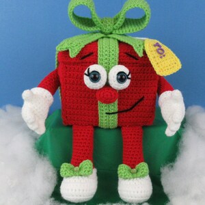 Puede incluir: Un juguete de peluche tejido a crochet con forma de caja de regalo con un lazo verde, un cuerpo rojo y piernas blancas. El juguete tiene una cara sonriente con ojos saltones y una cinta verde alrededor del medio. La caja de regalo tiene una etiqueta amarilla que dice "&iexcl;Para!"