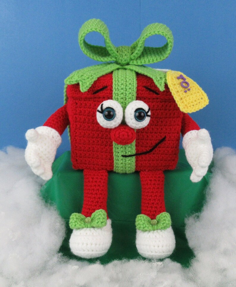 Puede incluir: Un juguete de peluche tejido a crochet con forma de caja de regalo con un lazo verde, un cuerpo rojo y piernas blancas. El juguete tiene una cara sonriente con ojos saltones y una cinta verde alrededor del medio. La caja de regalo tiene una etiqueta amarilla que dice "&iexcl;Para!"