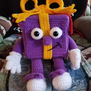 Puede incluir: Una caja de regalo de color morado tejida a crochet con un lazo amarillo y brazos y piernas blancos. La caja de regalo tiene una cara sonriente con ojos negros y una nariz amarilla.