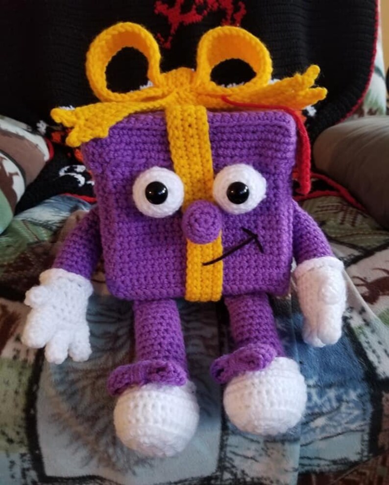Puede incluir: Una caja de regalo de color morado tejida a crochet con un lazo amarillo y brazos y piernas blancos. La caja de regalo tiene una cara sonriente con ojos negros y una nariz amarilla.