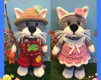 Könnte beinhalten: Zwei gehäkelte graue Katzenspielzeuge. Eine Katze trägt eine rote Latzhose mit einem Fischmuster und einen braunen Hut. Die andere Katze trägt ein rosa Kleid mit einer weißen Schleife und einer rosa Blume.