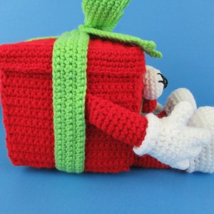 Puede incluir: Una caja de regalo roja de crochet con una cinta verde y brazos y piernas de crochet blanco.