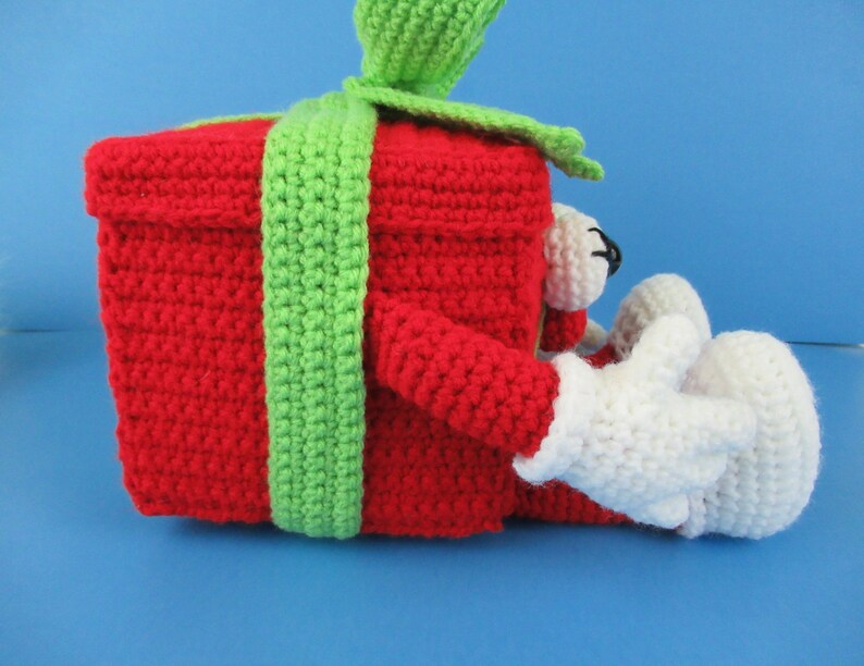 Puede incluir: Una caja de regalo roja de crochet con una cinta verde y brazos y piernas de crochet blanco.