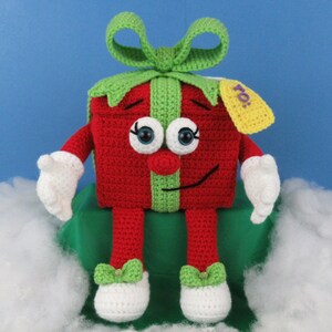 Puede incluir: Un juguete de peluche tejido a crochet con forma de caja de regalo con un lazo verde, un cuerpo rojo y brazos y piernas blancos. El juguete tiene una cara sonriente con ojos grandes y una nariz roja. La caja de regalo tiene una etiqueta verde con la palabra "To!" en ella.